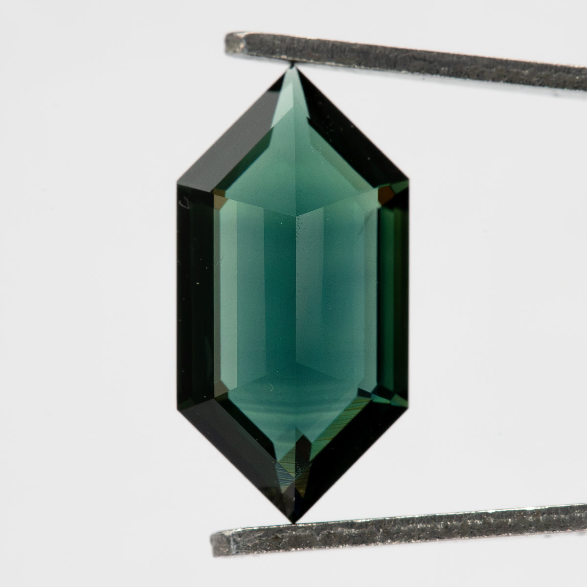 Pavilion view of the teal green-blue sapphire held upright in tweezers, showing its elongated six-sided outline and central keel.