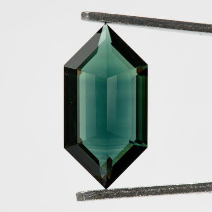Pavilion view of the teal green-blue sapphire held upright in tweezers, showing its elongated six-sided outline and central keel.