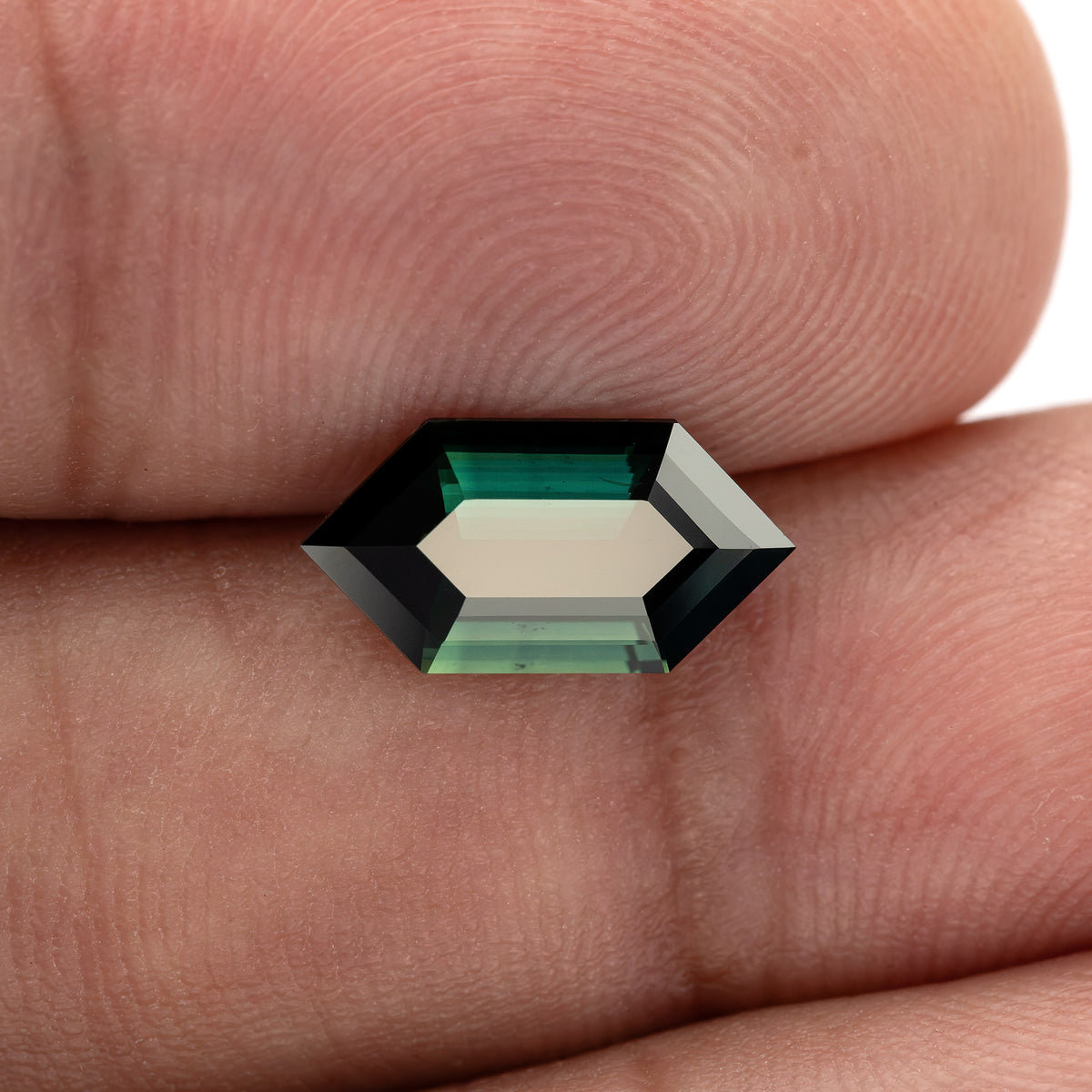 The elongated sapphire resting between fingers for scale, with a pale central reflection and green-blue bands along its long sides.