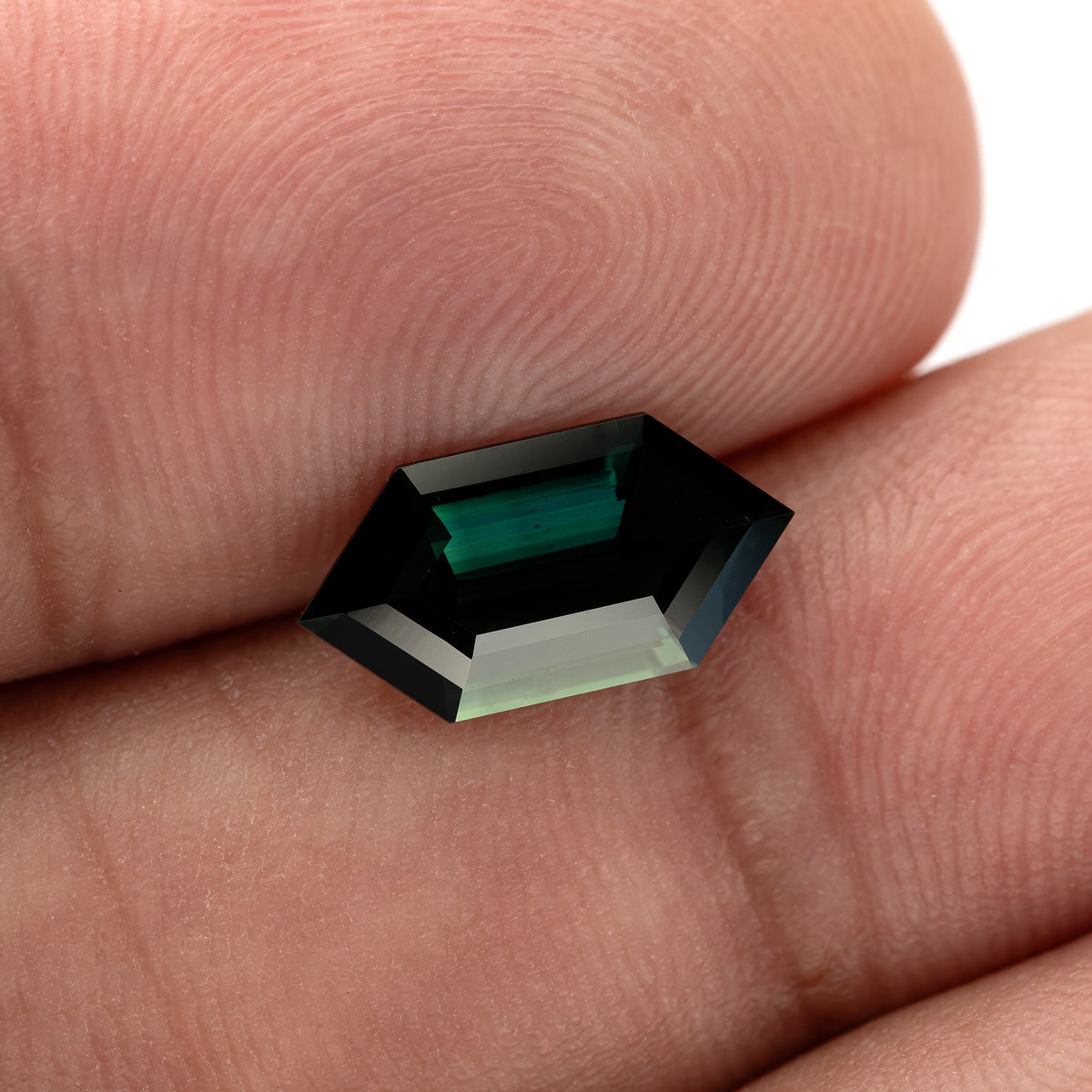 The sapphire tilted between fingers, showing the contrast between dark teal areas and a broad pale reflection.