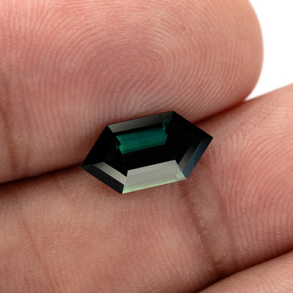 The sapphire tilted between fingers, showing the contrast between dark teal areas and a broad pale reflection.