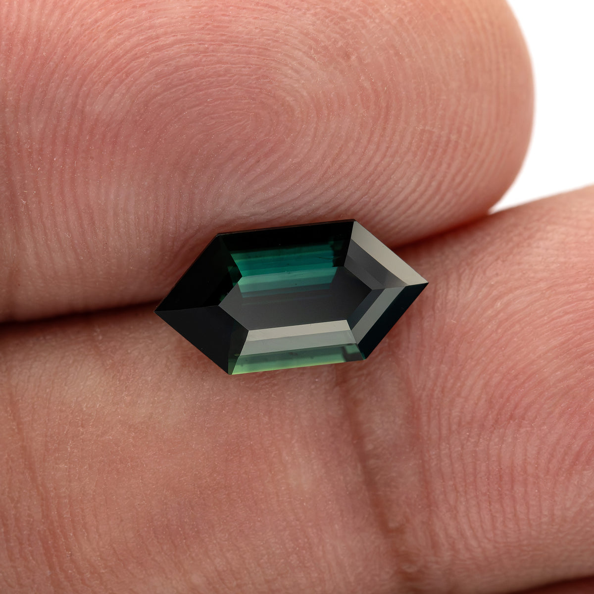 Loose green-blue sapphire held between fingers, showing its angular outline, stepped facets and real-life scale.