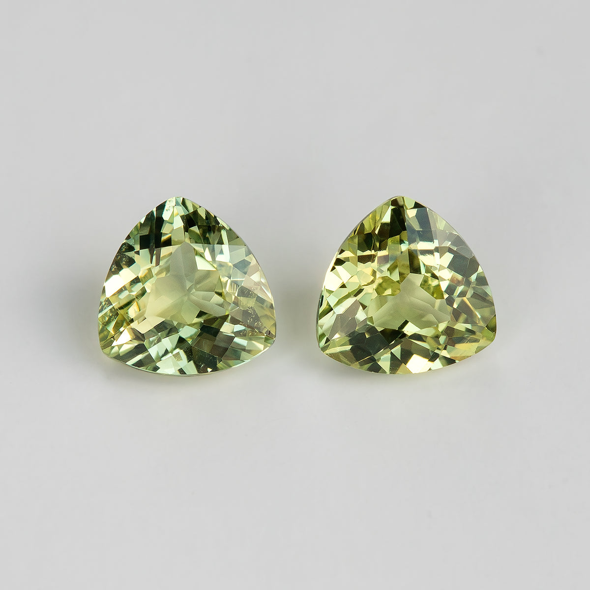 Face-up view of two yellow-green trillion sapphires on a white surface, showing complete rounded triangular outlines and olive-green and golden reflections.