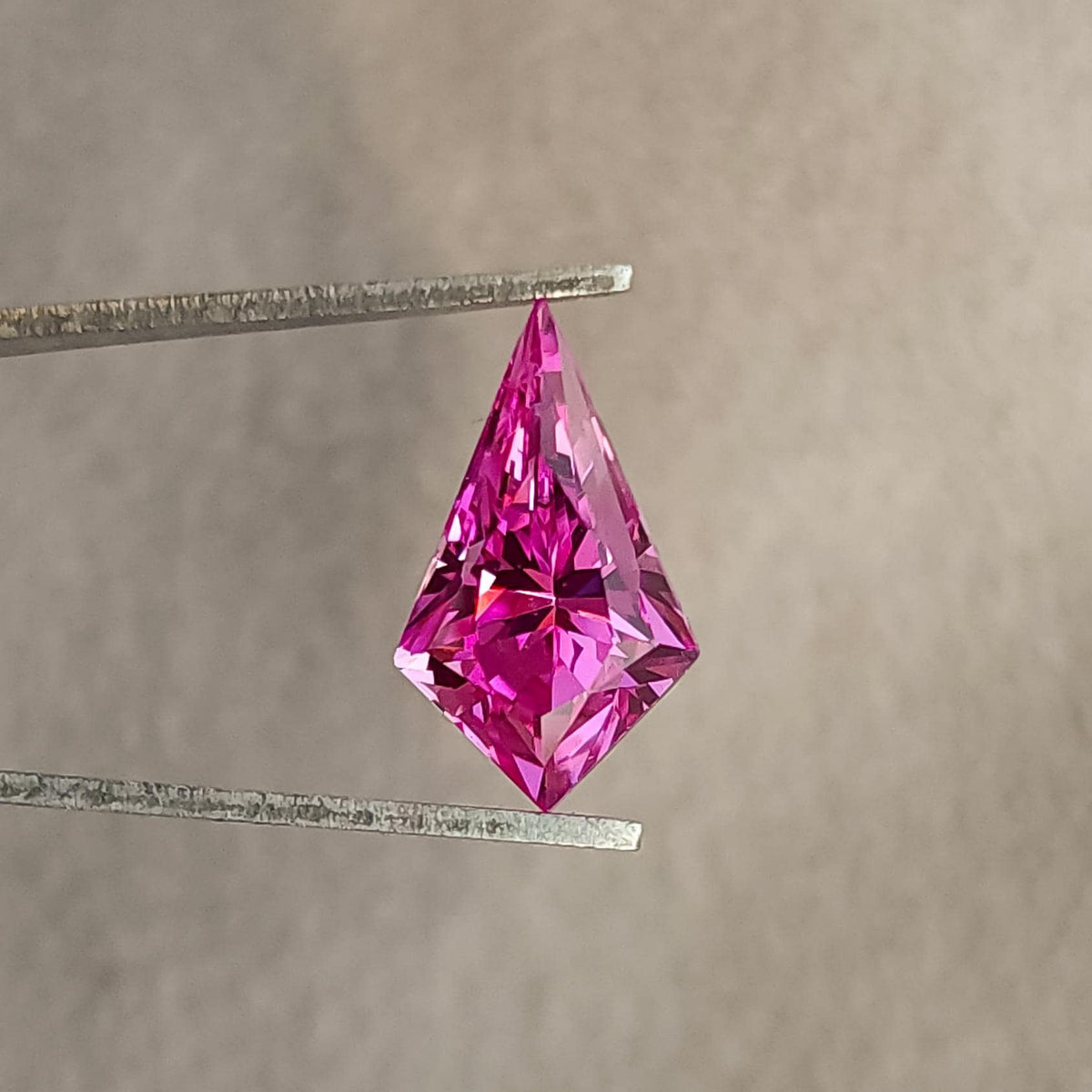 Vivid pink geometric kite cut gemstone held vertically in metal tweezers against a plain textured background.