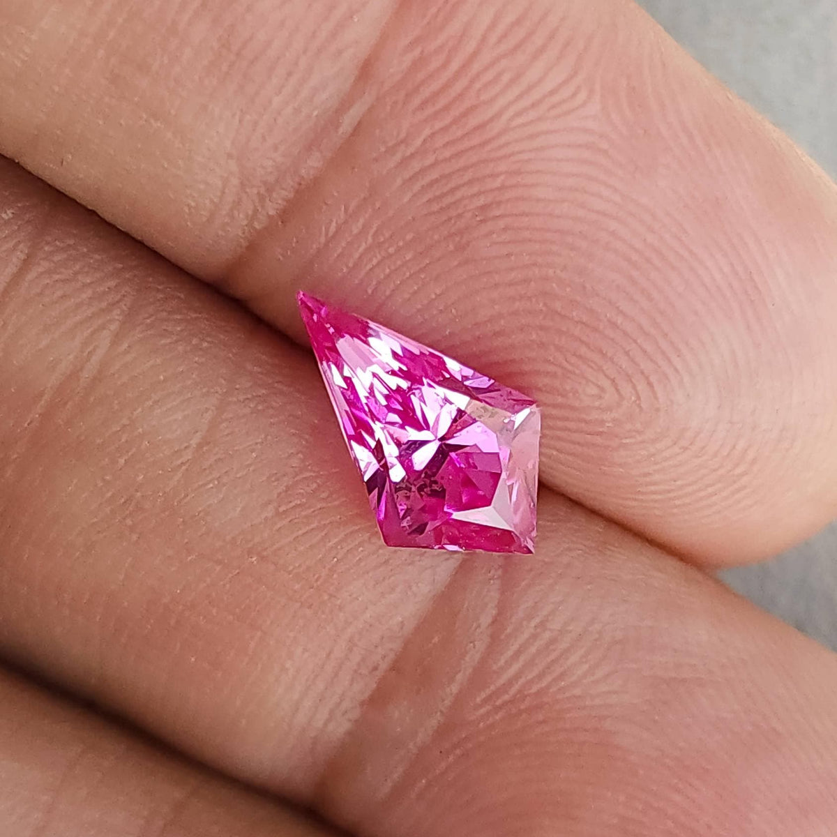 Geometric kite cut pink gem resting diagonally across human fingers, showing cut facets and color saturation.