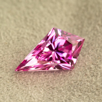 Close-up angled view of a vivid pink faceted geometric kite gemstone on soft grey fabric.