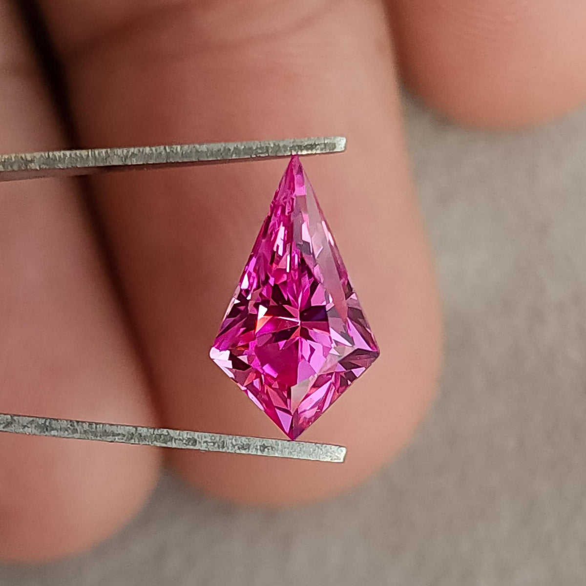 Face-up view of a bright pink geometric kite cut gemstone held in tweezers against fingers.