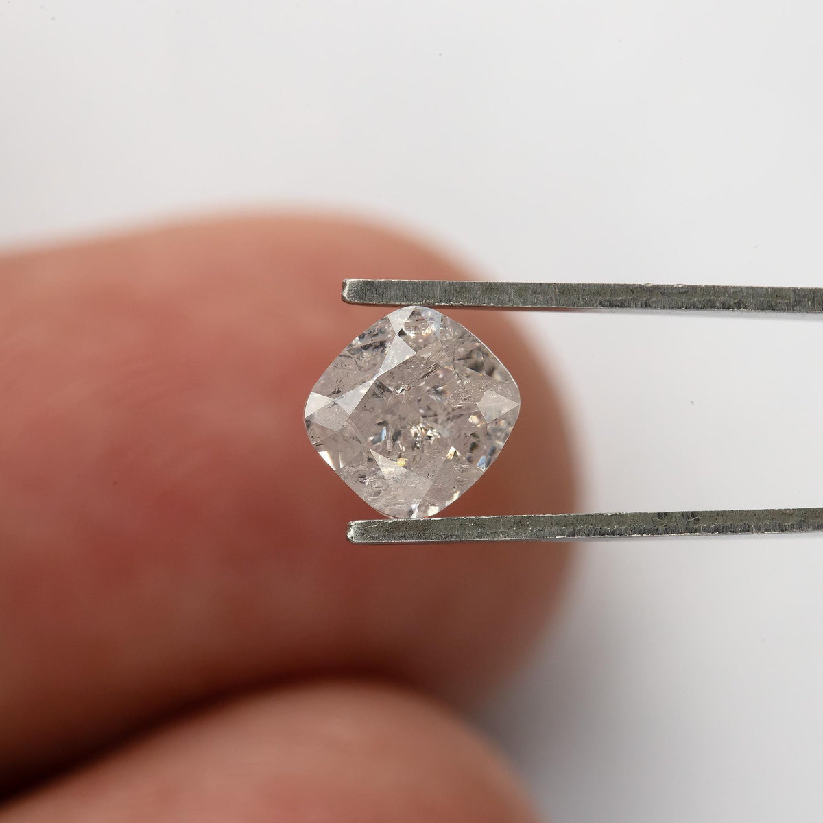 Light pink cushion diamond held face-up in tweezers against a fingertip for relative size.