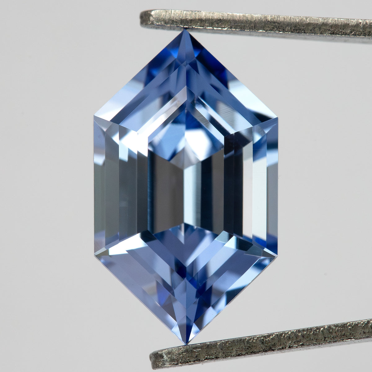 Face-up view of a cornflower-blue lab-grown sapphire between tweezers, showing its elongated hexagonal shape.