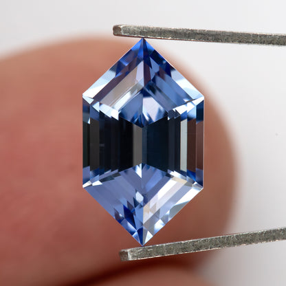 Cornflower-blue lab-grown sapphire held vertically between tweezers against a fingertip for scale.