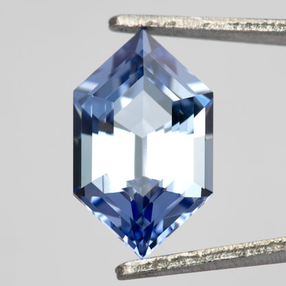 Cornflower-blue lab-grown sapphire between tweezers, showing a pale central area and blue reflections near the pointed ends.