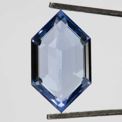 Cornflower-blue lab-grown sapphire between tweezers, showing a broad pale centre framed by blue edge facets.
