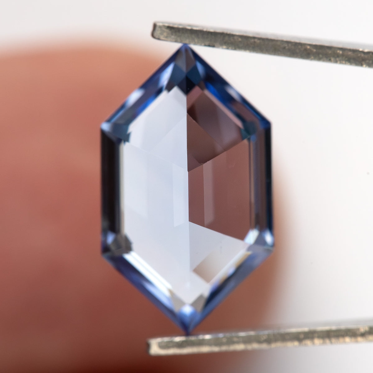 Cornflower-blue lab-grown sapphire between tweezers against a fingertip, showing a broad pale central area.