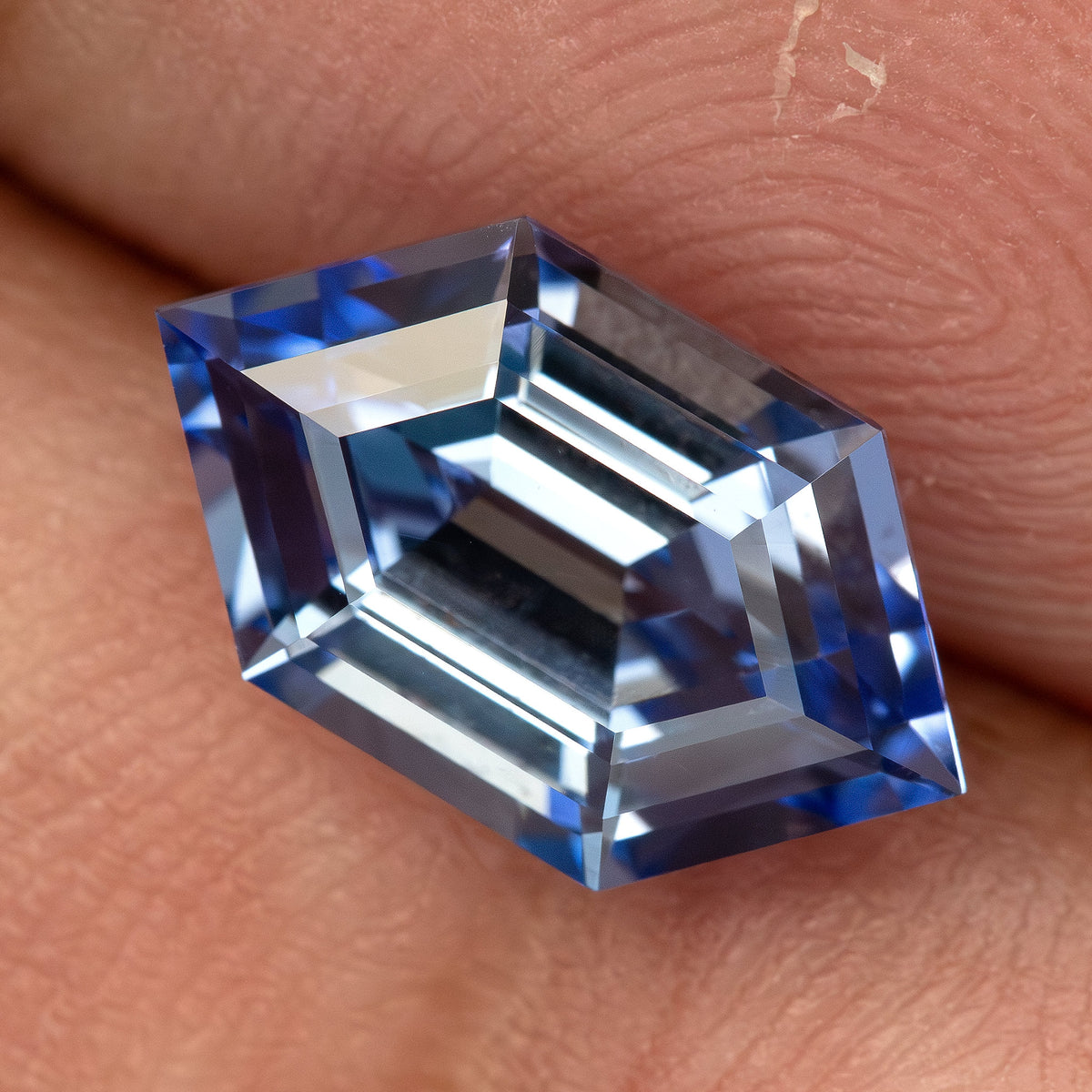 Angled close-up of a cornflower-blue lab-grown sapphire against fingers, showing dark blue and bright step facets.