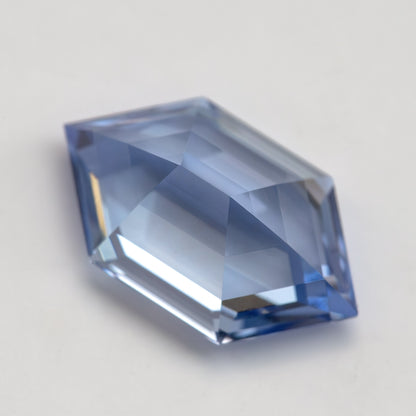 Low angled view of a cornflower-blue lab-grown sapphire on a pale surface, showing its profile and broad pale facets.