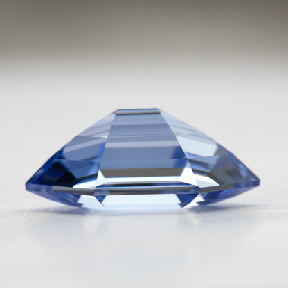 Side profile of a cornflower-blue lab-grown sapphire on a pale surface, showing its faceted outline.