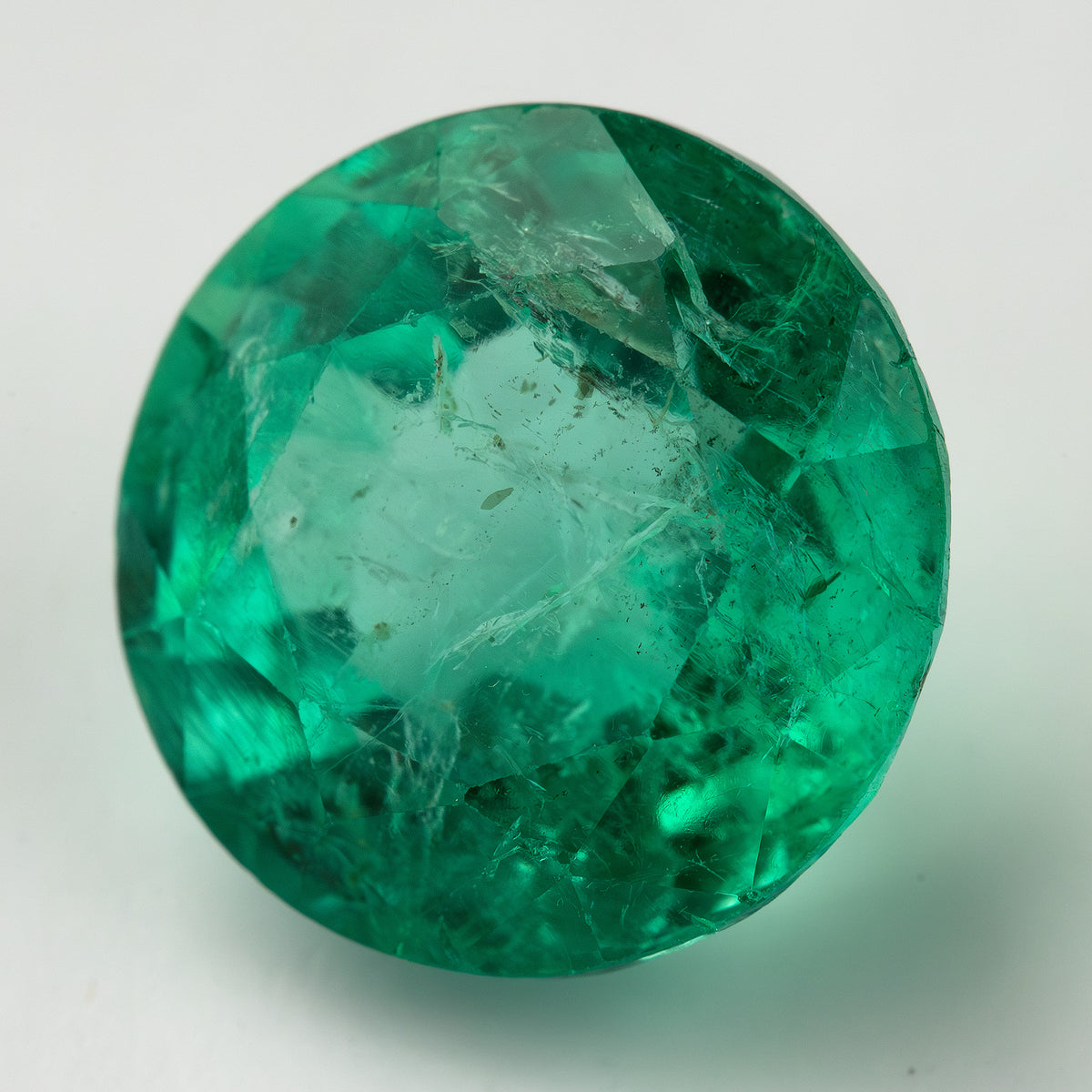 Large round green emerald photographed face-up on white, showing a pale central area and dense internal features around the rim.