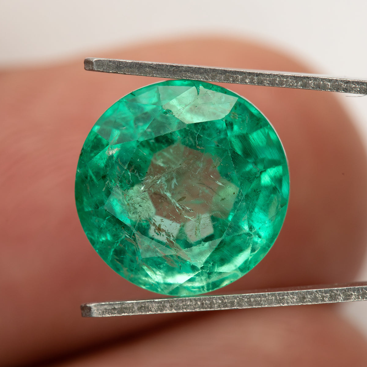 Face-up tweezer view of the round green emerald with a fingertip behind it for scale.