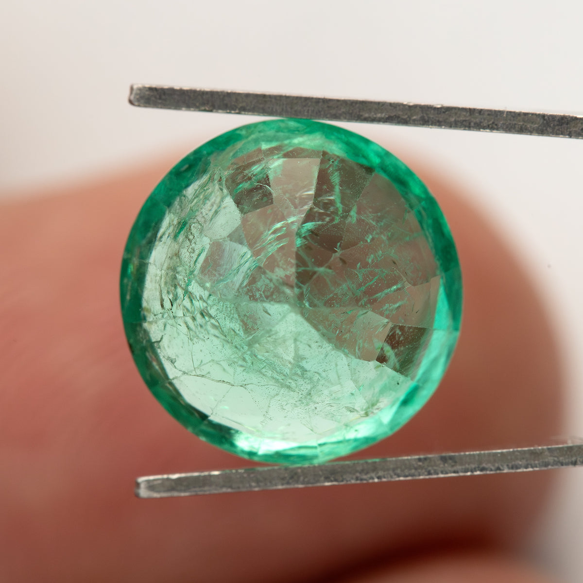 Angled face-up tweezer view of the round green emerald against a fingertip.