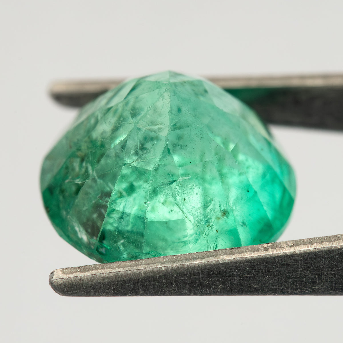 Side view of the round green emerald held in tweezers, with the pavilion extending below the girdle.