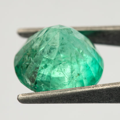Side view of the round green emerald held in tweezers, with the pavilion extending below the girdle.
