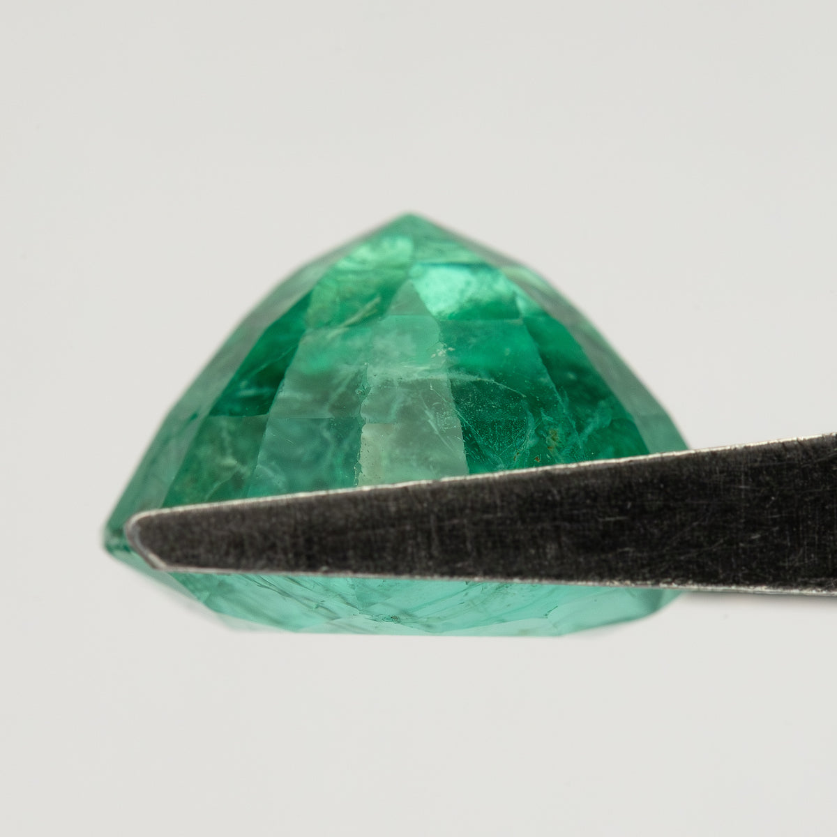 Low side view of the round green emerald in tweezers, showing pavilion depth and edge outline.