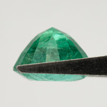 Low side view of the round green emerald in tweezers, showing pavilion depth and edge outline.