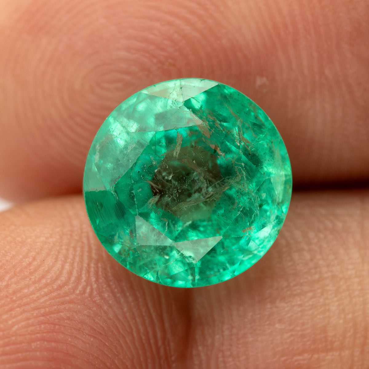 Round green emerald resting face-up on a fingertip for relative scale, with internal features visible through the center.