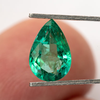 Pear emerald held face-up in tweezers against a blurred finger background.