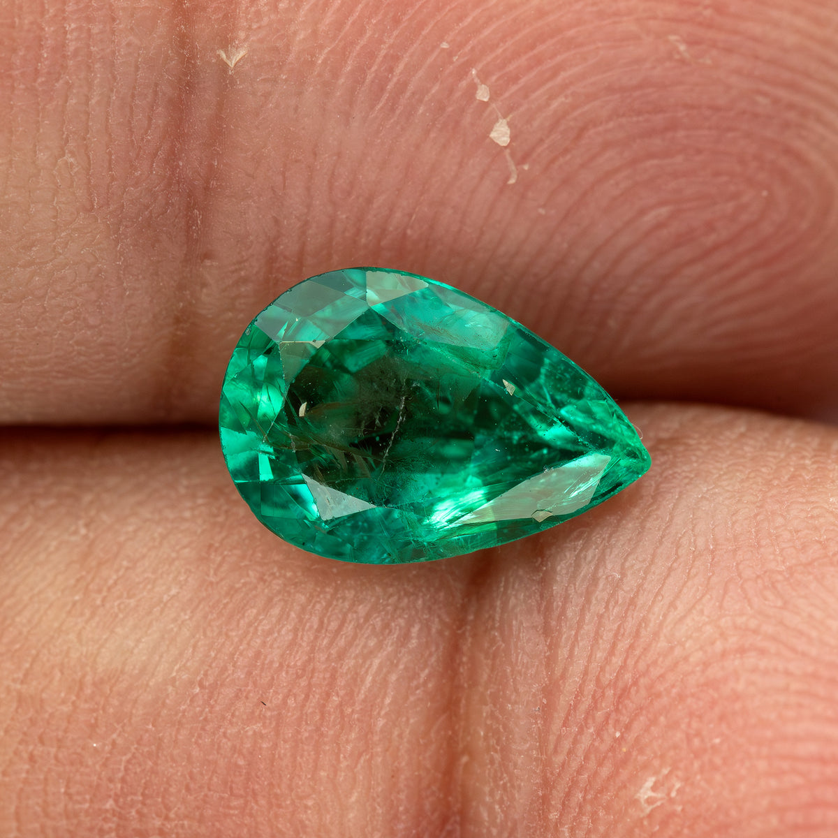 Pear emerald lying face-up horizontally between fingertips for scale.
