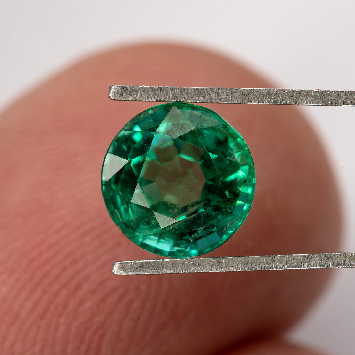 Round green emerald held face-up in tweezers with a fingertip behind it, showing the complete outline and visible internal features.