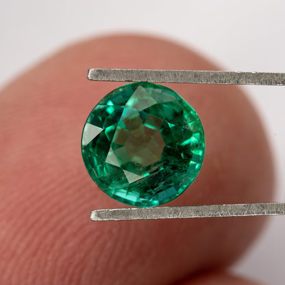 Round green emerald held face-up in tweezers with a fingertip behind it, showing the complete outline and visible internal features.