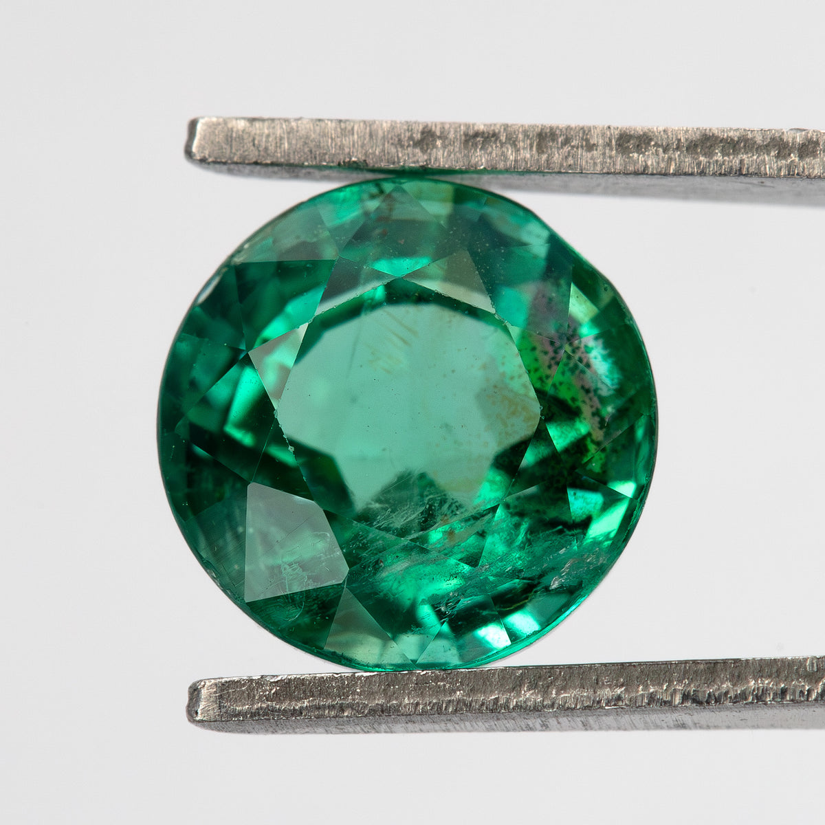 Second face-up tweezer view of the round green emerald on white, with a broad pale center and darker green rim.