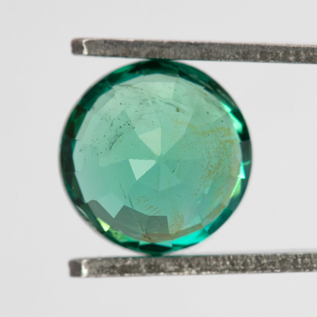 Close face-up table view of the round green emerald in tweezers, with fine internal features visible.