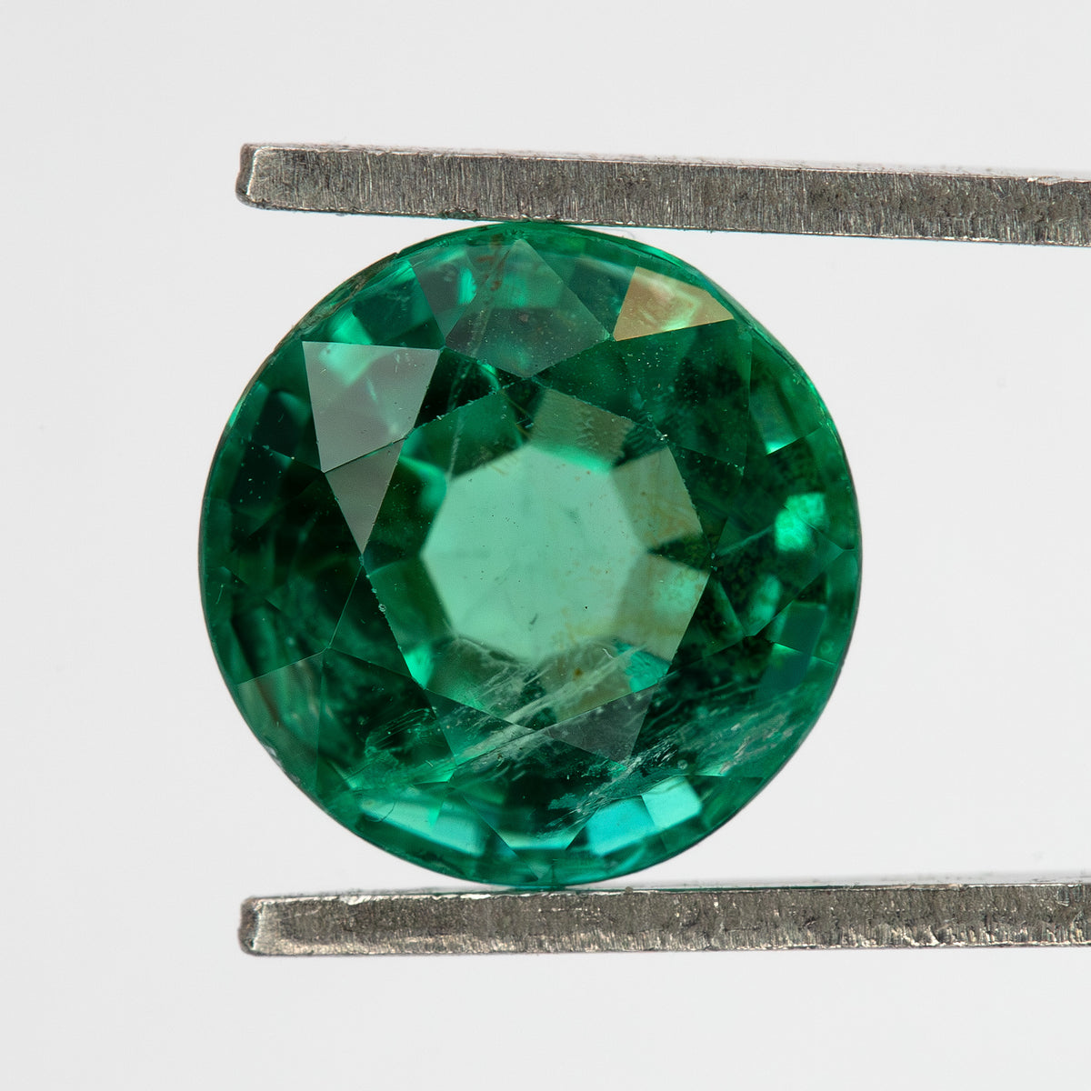 Face-up tweezer view of the round green emerald on white, showing a different central reflection.