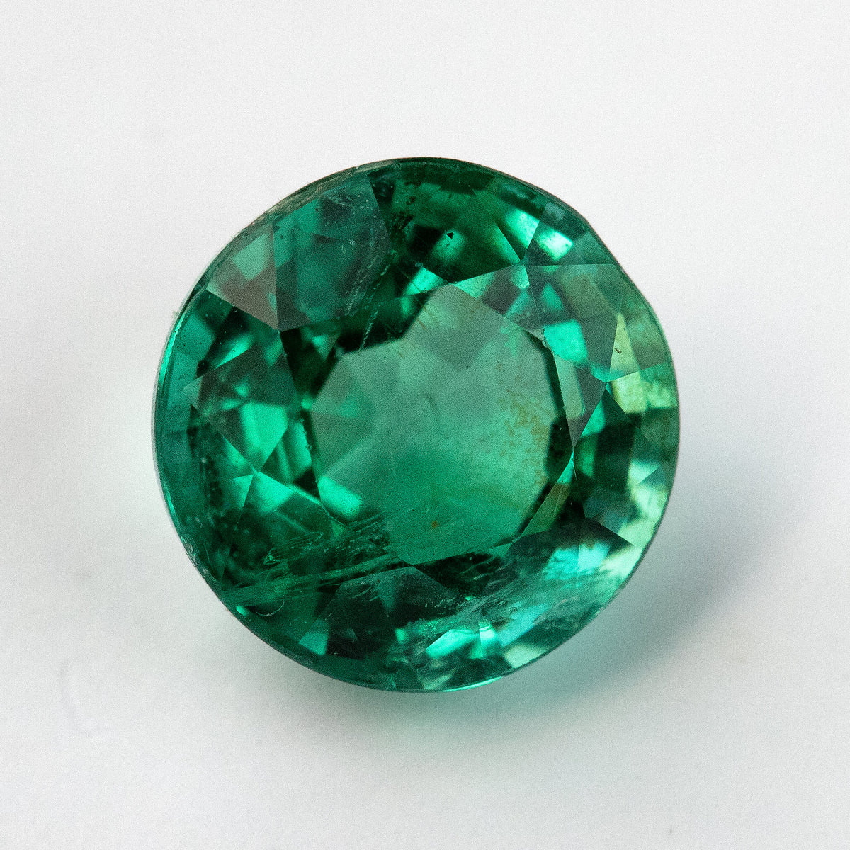 Third loose face-up view of the round green emerald on white, showing edge facets and internal features.