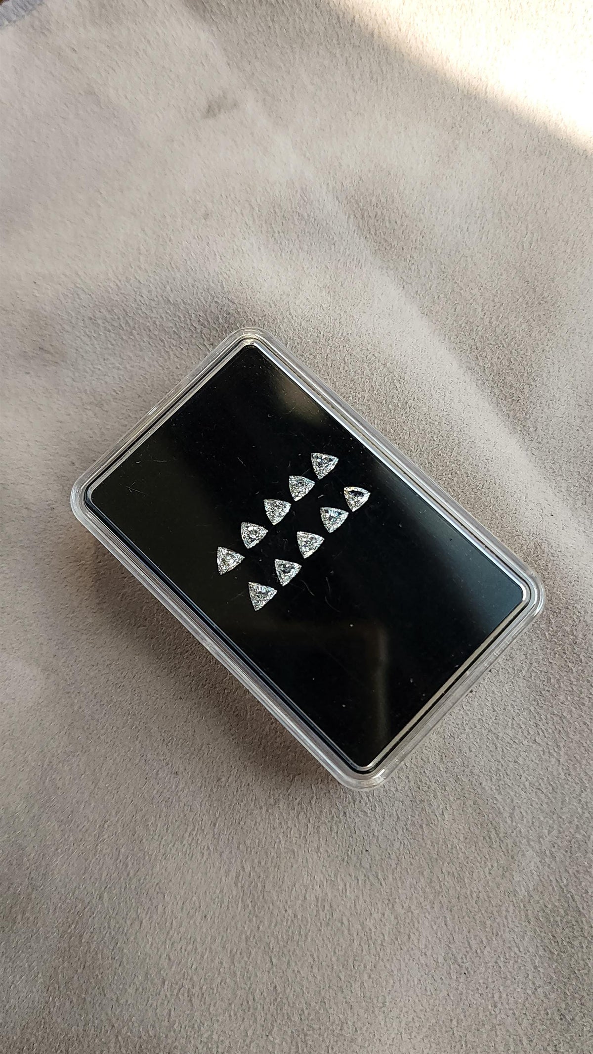 Ten loose trillion-cut diamonds on a black pad inside a clear case, resting diagonally on light textured fabric. Representative stock.