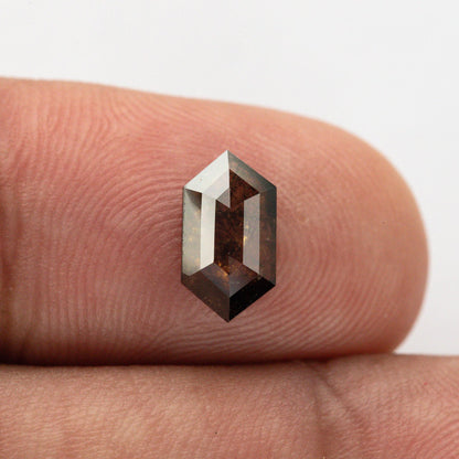 Opposite face of the elongated hexagon diamond on a fingertip, showing its second faceted surface and warm brown reflections.
