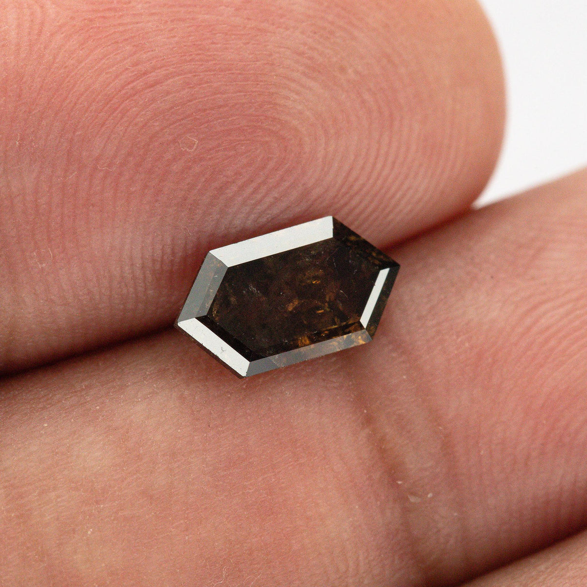 Geometric brown diamond resting flat on a fingertip, showing its elongated outline, pointed ends and dark angular facets.