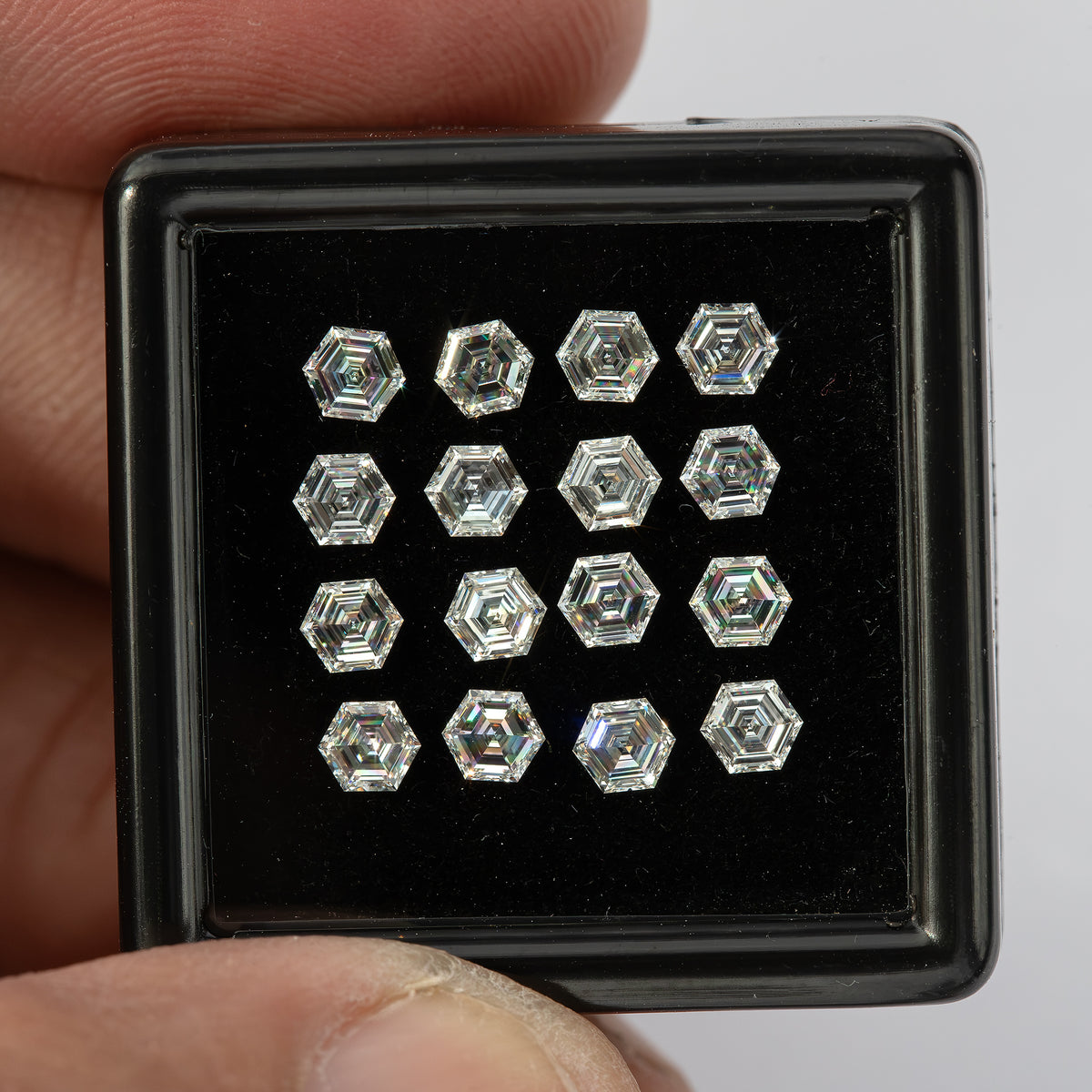 Face-up tray of hexagonal diamonds held near a fingertip, with bright perimeter facets and dark central reflections.