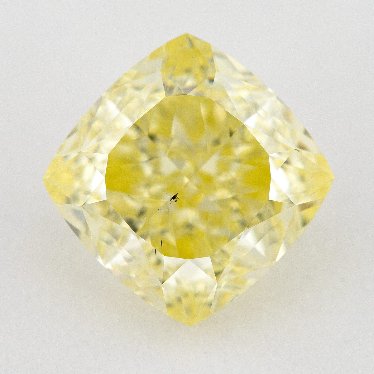 Yellow cushion-shaped natural diamond face-up on a pale surface, with dark internal characteristics visible near the center.