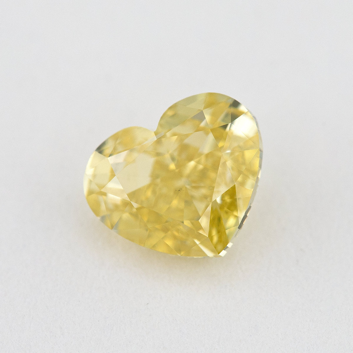 Yellow heart-shaped natural diamond tilted on a pale surface, showing the crown facets and pointed outline.