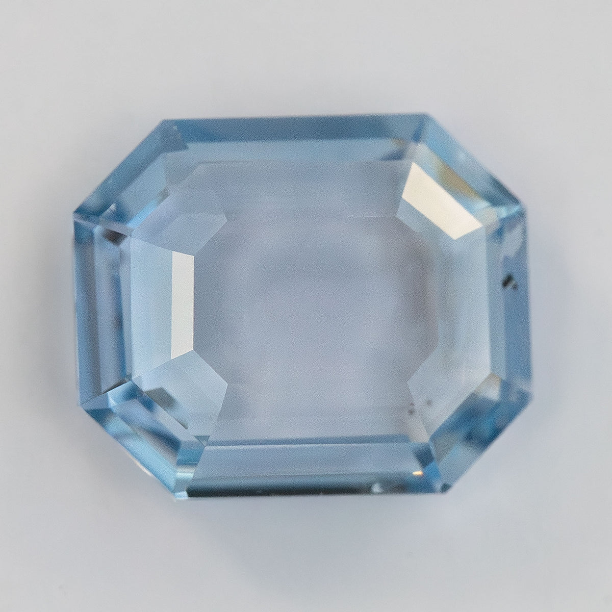 Octagonal portrait step cut light blue sapphire shown face-up on a plain white surface.