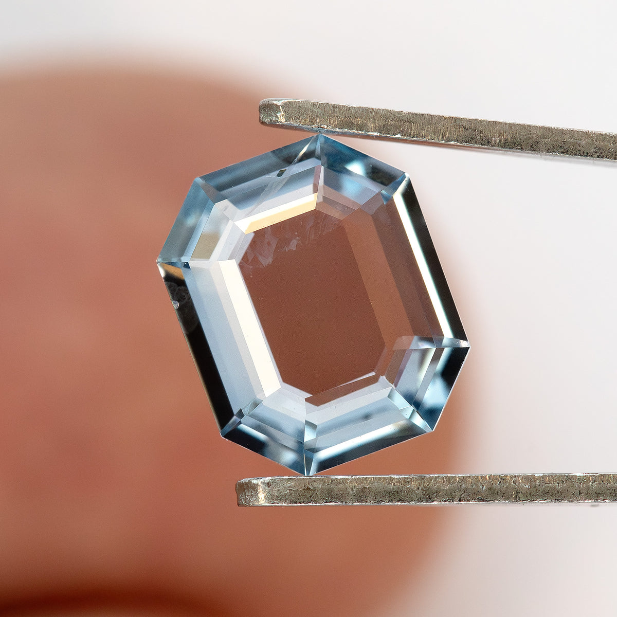 Light blue octagonal sapphire held in metal tweezers over a hand background.