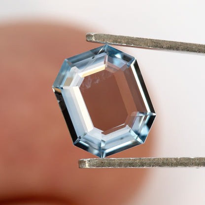 Light blue octagonal sapphire held in metal tweezers over a hand background.
