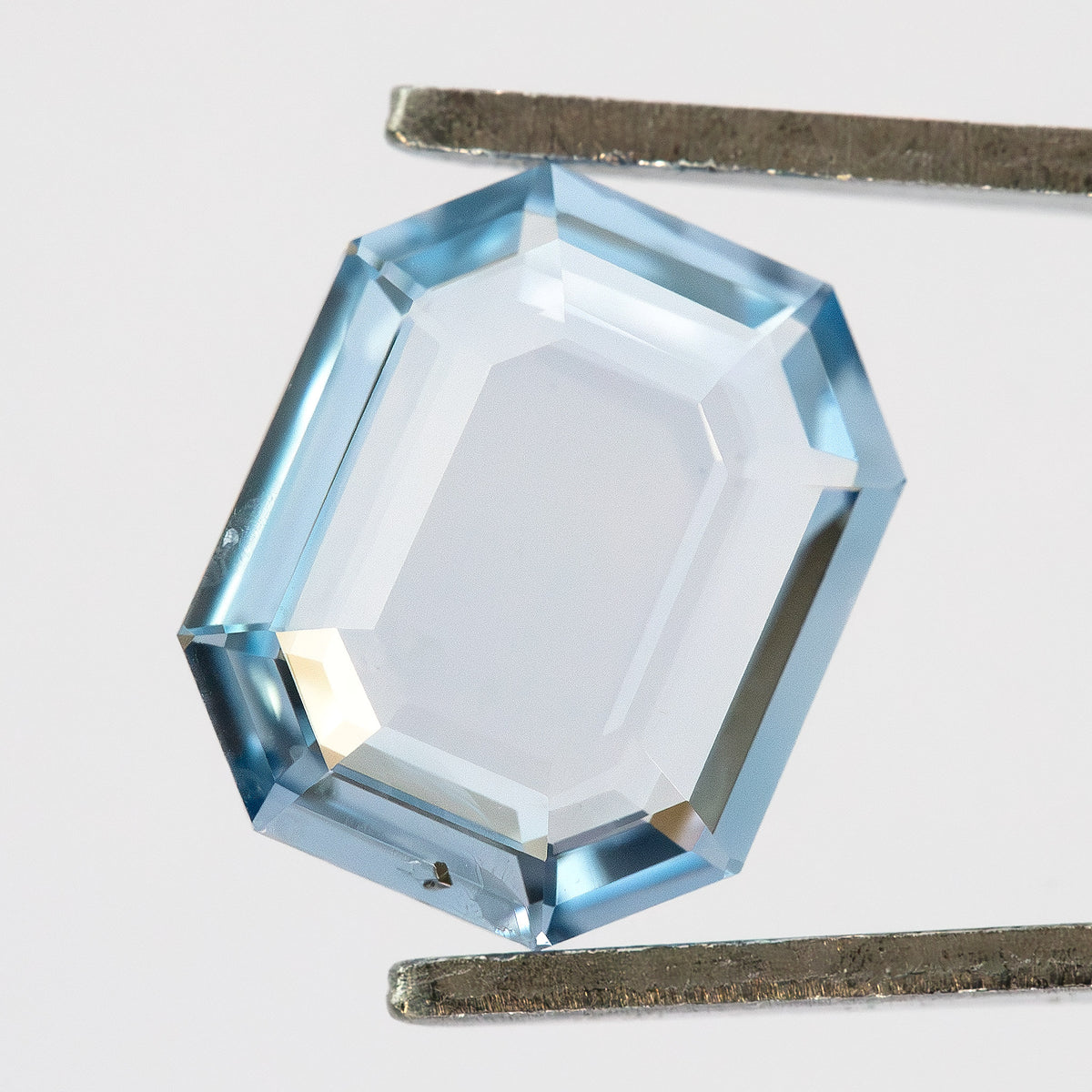 Light blue portrait step cut sapphire held horizontally in tweezers over a bright background.