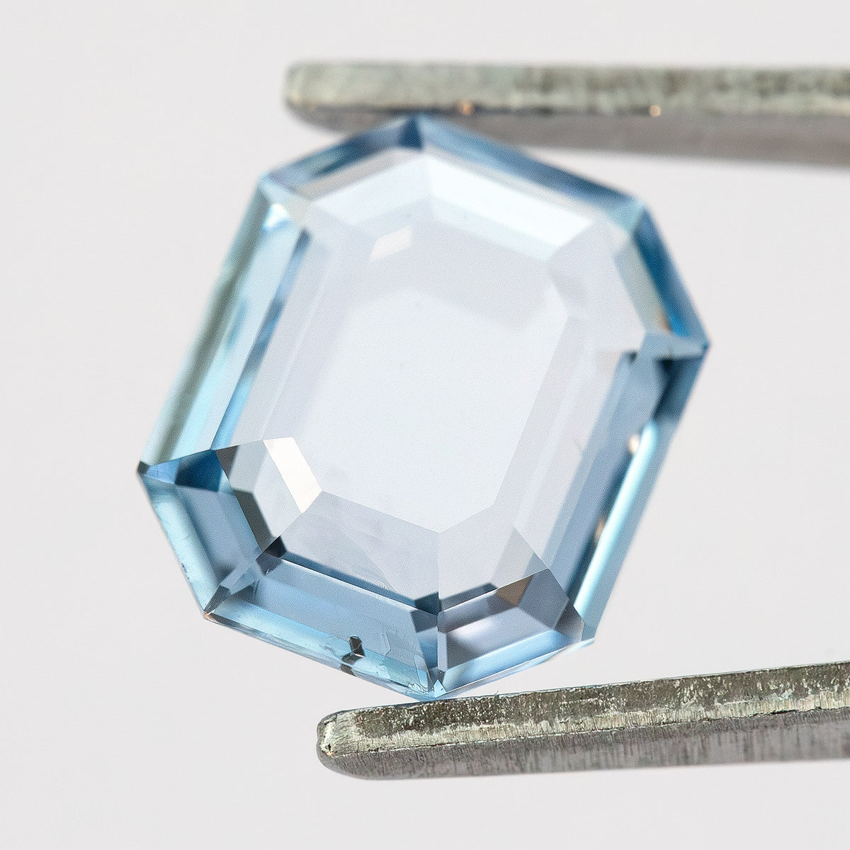 Tilted view of light blue octagonal step cut sapphire held in tweezers showing bright facet highlights.