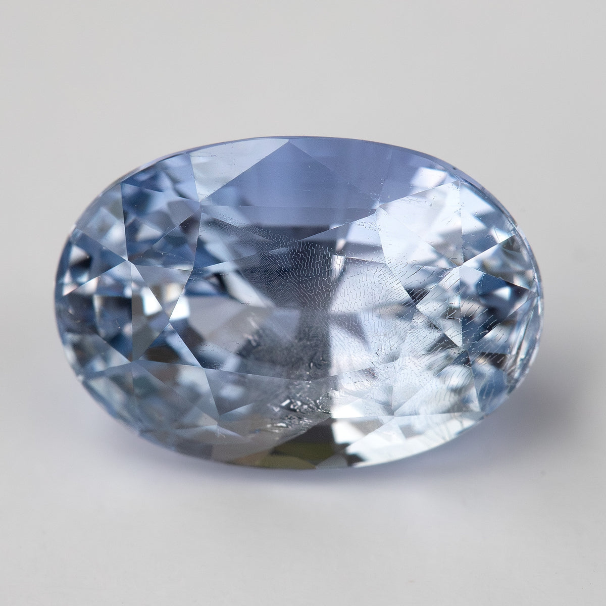 Close face-up view of the light blue oval sapphire on a pale background, showing its facet reflections and natural internal features.