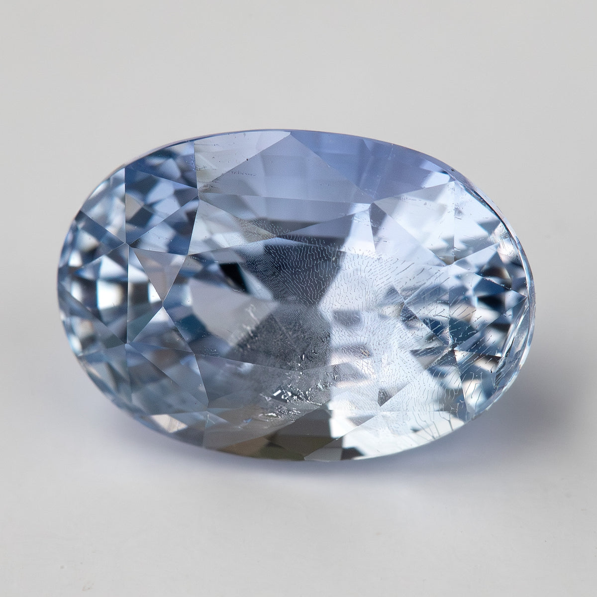 Direct face-up view of the light blue oval sapphire on a clean pale background, showing its crown facets and natural internal features.