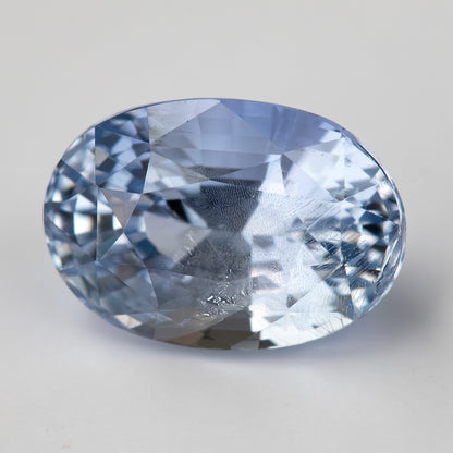 Direct face-up view of the light blue oval sapphire on a clean pale background, showing its crown facets and natural internal features.